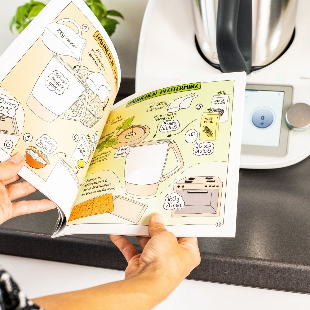Das aufgeschlagene Rezeptbuch mit dem Thermomix® im Hintergrund